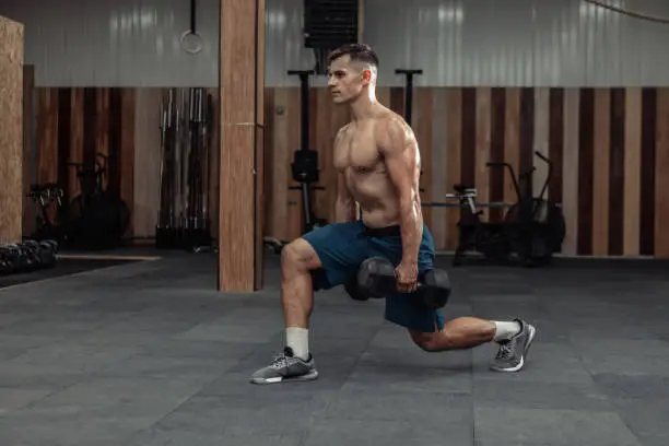 Photo of Muscular man training his legs, doing lunges with heavy dumbbells in modern gym. Healthy lifestyle concept