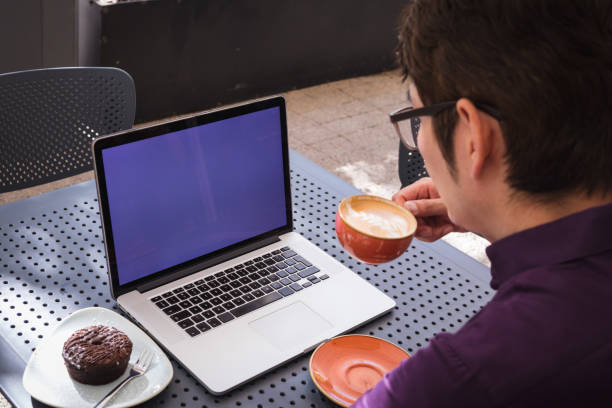 Asian businessman using laptop with blank screen drinking coffee in cafe. digital nomad out and about in city concept.