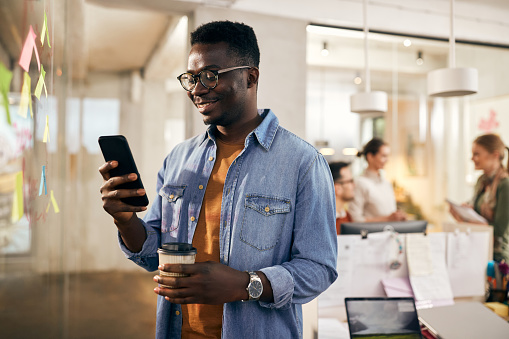 Happy black entrepreneur using smart phone while having coffee break in the office. His colleagues are in the background.