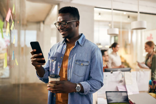 Young African American freelancer text messaging on mobile phone during coffee break at casual office. Happy black entrepreneur using smart phone while having coffee break in the office. His colleagues are in the background. coffee break photos stock pictures, royalty-free photos & images
