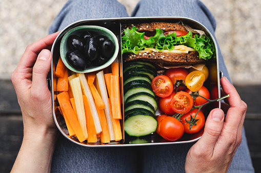 Point of view of a woman having a healthy lunch box. Black olives, carrots, cucumber, Cherry tomatoes and sandwich in the lunch box.