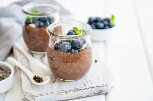 Chocolate chia pudding with blueberry, almonds and mint on top in a glass jar on a white wooden background. Healthy food. Copy space