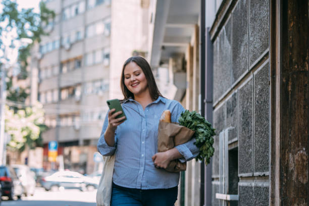 istock Smiling  plus size Woman Using her Smartphone while Walking Home from the Marketplace, Carrying a Reusable Bag 1321204080