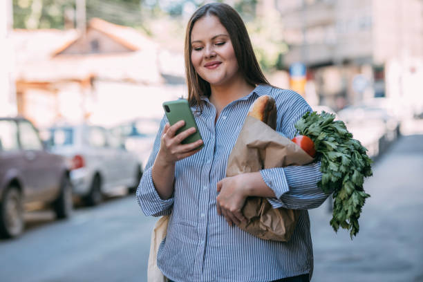 istock Portrait of a Smiling  Plus Size  Woman Using her Smartphone while Walking Home from the Marketplace, Carrying a Reusable Bag 1321204642