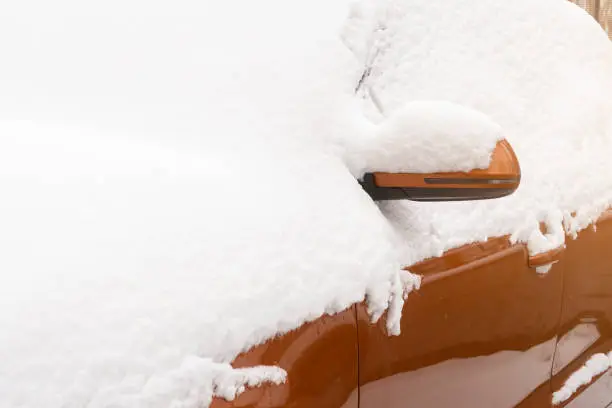 Orange car covered with fresh white snow. View after snowfall in winter season. Mirrow and side are partially visible.