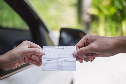 Close-up shot of unrecognizable patient in a car receiving a COVID-19 vaccination record card at a drive through center from a healthcare worker.