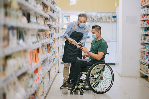 asian indian man with disability on wheelchair asking senior man retail assistant on his shopping list from smart phone at supermarket during weekend