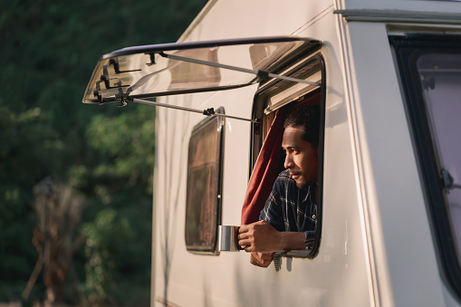 Handsome young Asian man, traveler on road trip, sit inside camping van in the morning. Cosy comfortable setup in camper trailer or van. Millennial travel trend, adventure on the road.