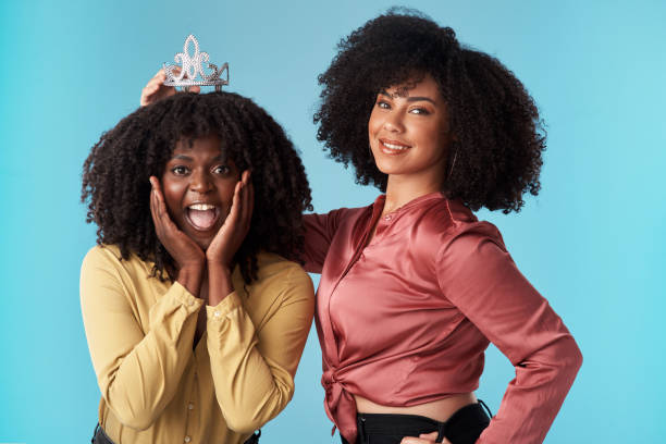 Studio shot of a young woman putting a crown on her friend against a blue background We're all winners when we support each other beauty queen stock pictures, royalty-free photos & images