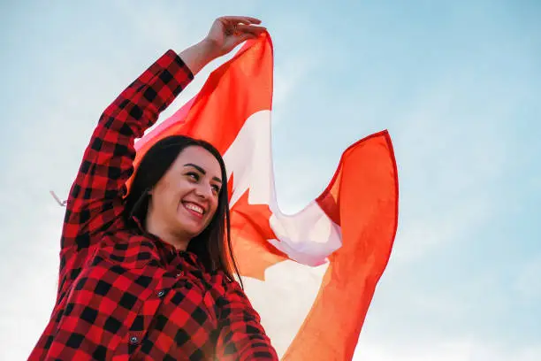 Young millennial brunette woman holding The National Flag of Canada. Canadian Flag or the Maple Leaf. Tourist traveler or patriotism. Immigrant in a free country. Independence day 1th july. Canada day, copy space. National holiday Flag blowing in the wind