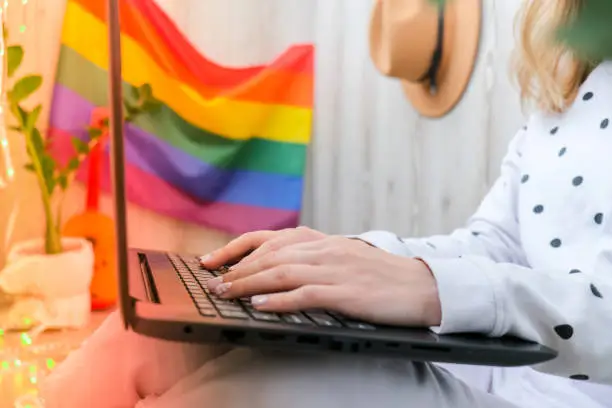 Photo of Young millennial hippie woman sitting on balcony using laptop. LGBTQ rainbow flag on background. Online education tuition. Work from home. Modern office