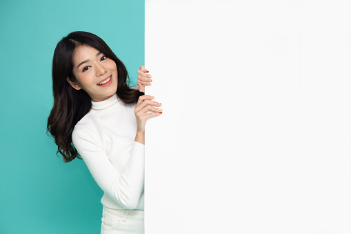 Cheerful Young Asian woman is standing behind the white blank banner or empty copy space advertisement board on green background, Looking at camera