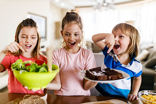 Little girl holding fresh salad and sweet cookies while her siblings choosing their favorite food.
