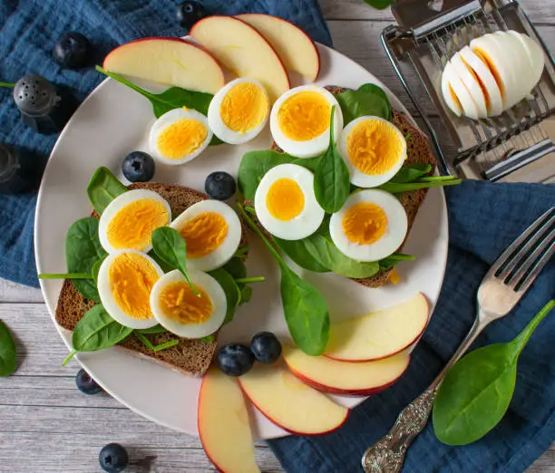 Egg Sandwich made with rye bread and boiled sliced eggs served with fresh spinach leaves and fruits on a plate. Overhead and closeup view