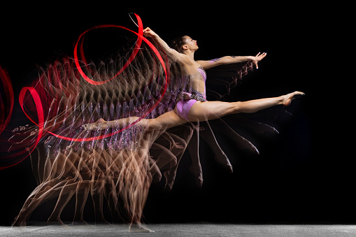 Flying with red ribbon. Young graceful girl, female rhythmic gymnast training isolated over dark background in mixed neon light. Concept of action, motion, sport life, competition. Copy space for ad.
