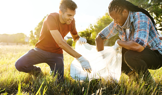 Two smiling diverse young male volunteers putting trash in garbage bags during a community cleanup day in a field