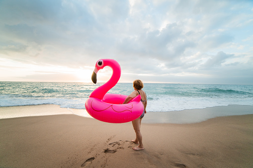 Senior woman standing on the sand beach with inflatable flamingo.