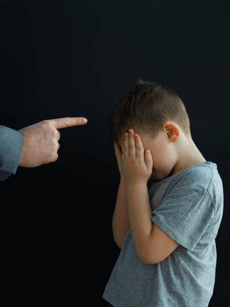 un uomo adulto punta severamente il dito indice contro un ragazzino che si trova di fronte a lui con le mani che gli coprono il viso - bullying family violence domination foto e immagini stock