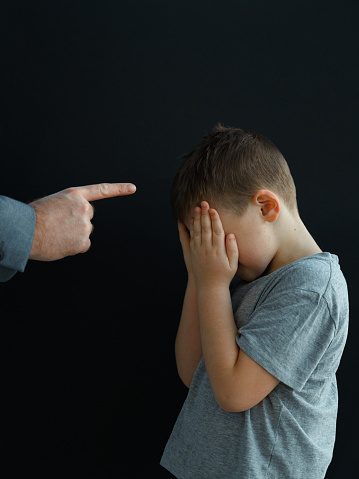 An adult man sternly points his index finger at a small boy who is standing in front of him with his hands covering his face. Perhaps the parent explains to the child that he behaved badly and will now be punished.