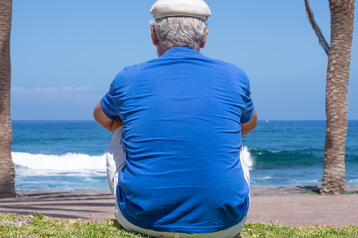 Back view of pensive senior man at beach looking away. Retired enjoying summer holiday at sea