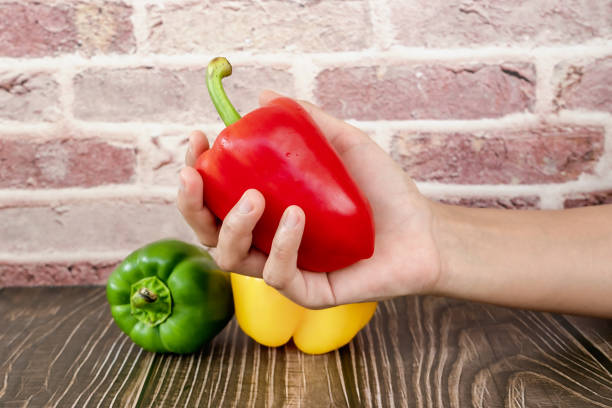 persona que sostiene pimiento rojo dulce en la mano con pimienta verde y amarilla en una mesa de madera con fondo de ladrillo. mano de mujer sosteniendo pimiento rojo fresco en verdura orgánica del jardín. alimentación saludable - green bell pepper bell pepper red bell pepper groceries fotografías e imágenes de stock