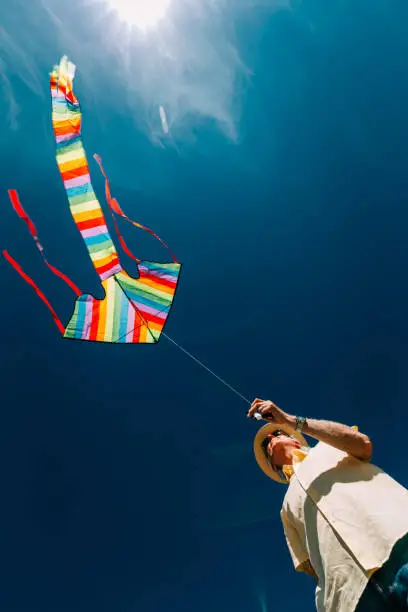 A senior man in his 60s flying a colorful rainbow kite at the beach on a hot summer day. The man is wearing casual clothing - a short-sleeved yellow shirt and denim jeans, with straw hat and sunglasses.