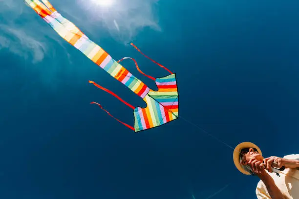 A senior man in his 60s flying a colorful rainbow kite at the beach on a hot summer day. The man is wearing casual clothing - a short-sleeved yellow shirt and denim jeans, with straw hat and sunglasses.