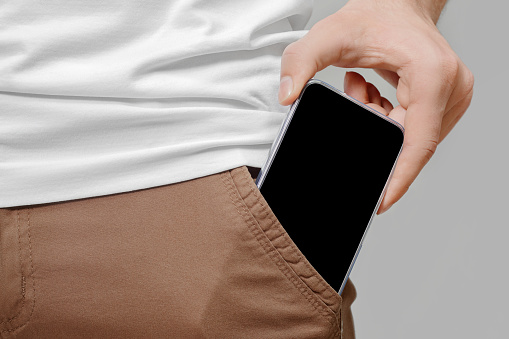 Close up of male hand taking out big mobile phone with empty black screen from front pocket of brown pants on grey background.