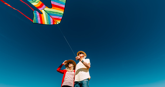 A senior couple in their 60s and 70s flying a colorful rainbow kite at the beach on a hot summer day. The man is wearing casual clothing - a short-sleeved yellow shirt and denim jeans, with straw hat and sunglasses, while the woman is also wearing a straw hat and red blouse.