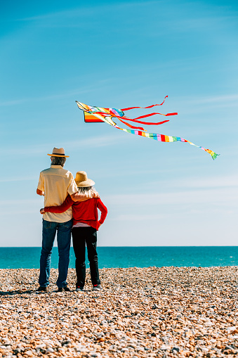 A senior couple in their 60s and 70s flying a colorful rainbow kite at the beach on a hot summer day. The man is wearing casual clothing - a short-sleeved yellow shirt and denim jeans, with straw hat and sunglasses, while the woman is also wearing a straw hat and red blouse.