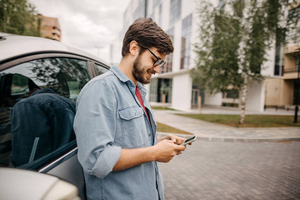 Young man using phone while waiting for friend Young man using phone in front of the car car insurance photos stock pictures, royalty-free photos & images