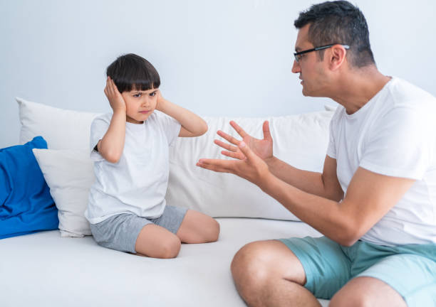 istock Irritated Father Shouting At Unhappy Little Son While He Covering Ears Sitting On Sofa At Home. Angry strict father shouting screaming at little son. 1321311470