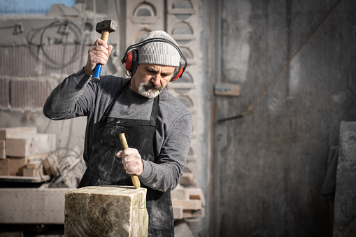 Stonemason wearing ear defenders while chiselling block of stone with chisel and hammer in workshop.