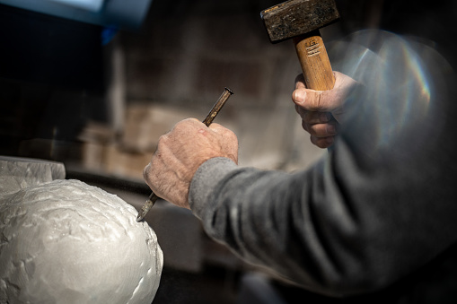 Close-up of stonemason carving a sculpture with chisel and hammer in the workshop.