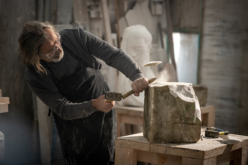 Stonemason wearing safety glasses while chiselling block of stone with chisel and hammer in workshop.