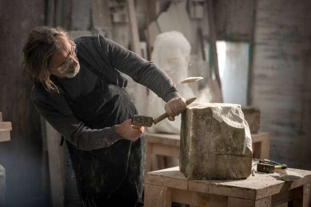 Stonemason chiselling a block of stone in workshop Stonemason wearing safety glasses while chiselling block of stone with chisel and hammer in workshop. sculptor stock pictures, royalty-free photos & images