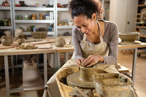 Smiling mature female potter making ceramic vase on a potter's wheel in workshop.