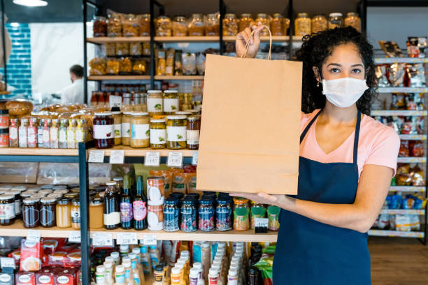 Owner with paper bag in store during COVID-19 Portrait of female store owner showing paper bag. Saleswoman is wearing protective face mask while standing in store. She is working during pandemic. convenience store stock pictures, royalty-free photos & images