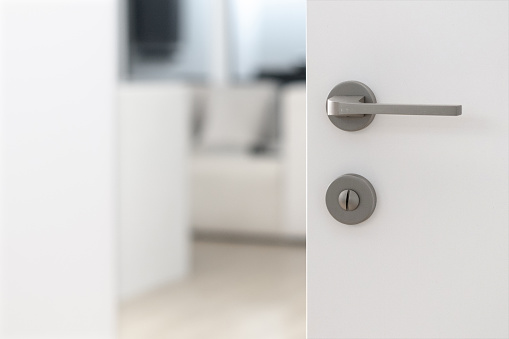 Closeup of slightly open white door with simple steel matt door handle and safety lock, blurred view of hotel room with modern white stylish sofa