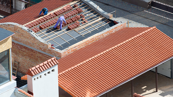 Concept of house, building under construction. High angle view of two professional roofers standing on new roof, install red ceramic tile on wooden frame with protective, waterproof layer at rooftop