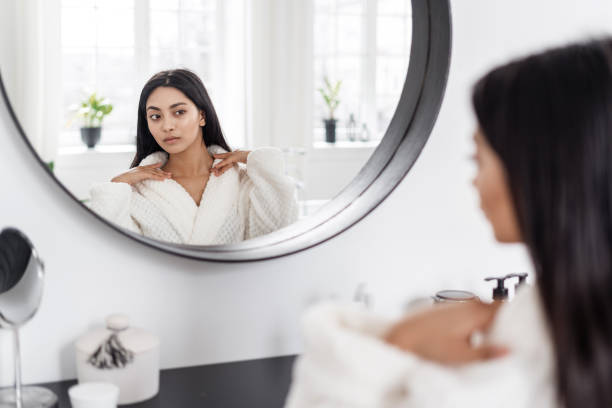 Young asian woman sitting in front of mirror looking at her skin Young good looking asian woman in white bathrobe looking at her neck, checking how smooth skin is while sitting in front of big mirror in bathroom at her flat. Female enjoying morning beauty routine neck photos stock pictures, royalty-free photos & images