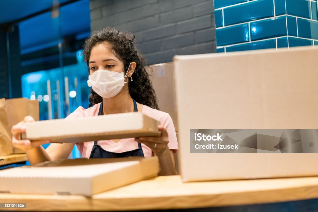 Saleswoman with pizza box in store during COVID-19 Saleswoman holding pizza box at counter. Female is wearing protective face mask while working. She is in store during COVID-19 crisis. Box - Container Stock Photo
