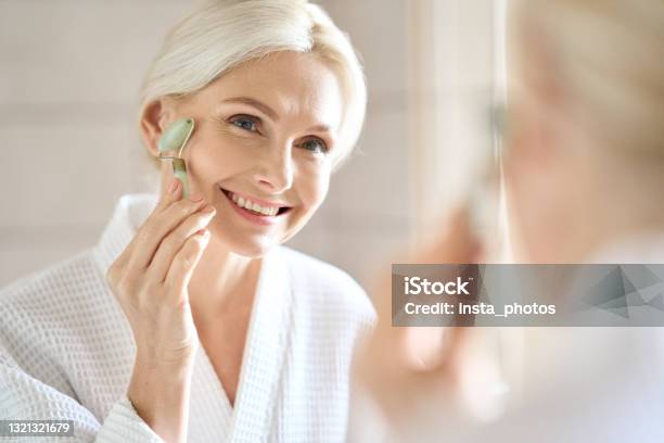 Senior Middle Aged Lady Massage Face Using Jade Stone Roller Looking At Mirror Stock Photo - Download Image Now