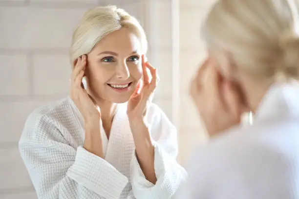 Photo of Attractive mid age woman smiling looking at mirror. Skincare concept.
