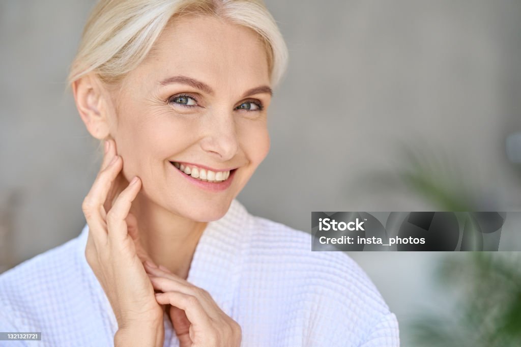 Portrait of smiling mid age woman looking at camera. Skin care concept. Closeup of happy smiling beautiful middle aged woman spa salon client wearing bathrobe looking at camera touching face. Spa procedures advertising. Skin care products concept. Copy space. Women Stock Photo