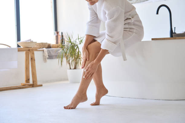 femme d’âge moyen d’âge mûr appliquant des veines crã ̈me sur les jambes assises dans la salle de bain. - jambe photos et images de collection