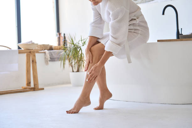 Middle aged mature woman applying veins crÃ¨me on legs sitting in bathroom. Mid age adult 50s age mature woman applying varicose prevention treatment cream massaging legs sitting on bathtub wearing white bathrobe. Feminine health care after menopause concept. blood flow stock pictures, royalty-free photos & images