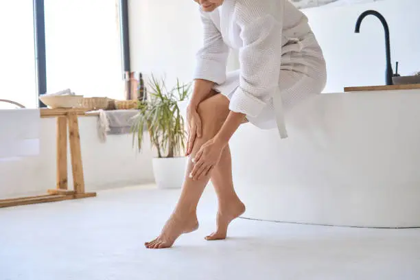 Mid age adult 50s age mature woman applying varicose prevention treatment cream massaging legs sitting on bathtub wearing white bathrobe. Feminine health care after menopause concept.