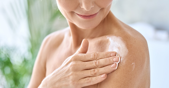 Naked middle aged 50s woman pampering herself doing daily routine applying nourishing smoothing body sun cream lotion after shower. Advertising of sunblock treatment. Closeup cutout.