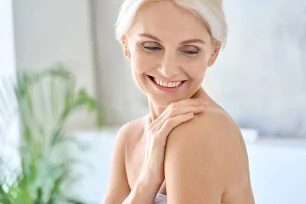 Portrait of happy smiling attractive senior adult nude blonde lady wearing white towel after morning shower touching embracing shoulder with pleasure. Body skin care concept.